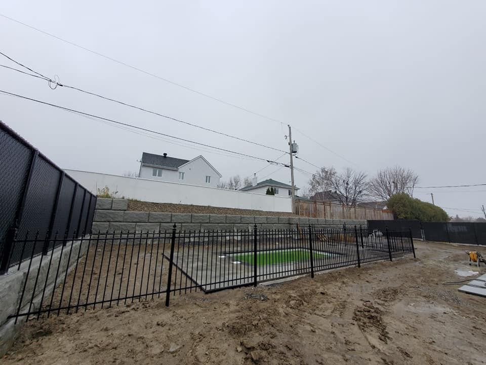 Une clГґture noire entoure un champ de terre avec une maison en arriГЁre-plan.